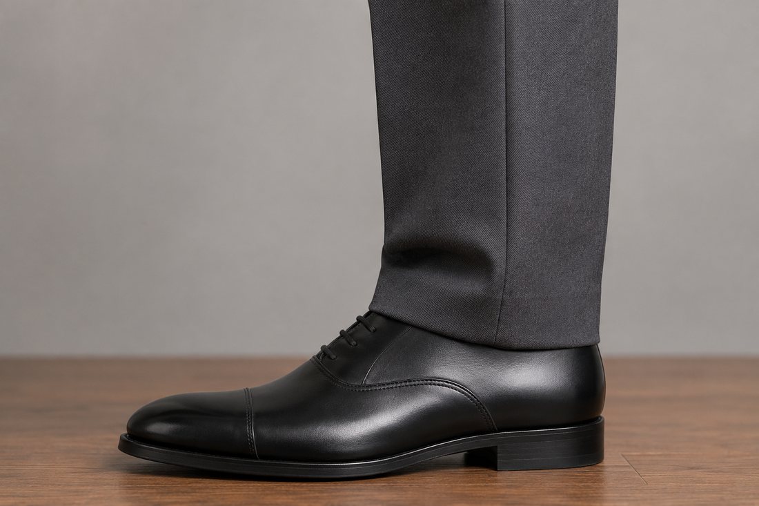 A charcoal trouser hem resting on a polished black Oxford shoe with one soft horizontal fold, covering two-thirds of the shoe