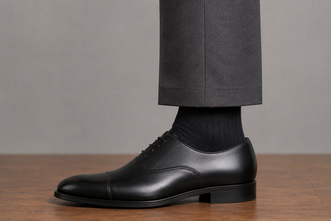 A charcoal trouser hem stopping well above the shoe, exposing a band of dark sock while standing still