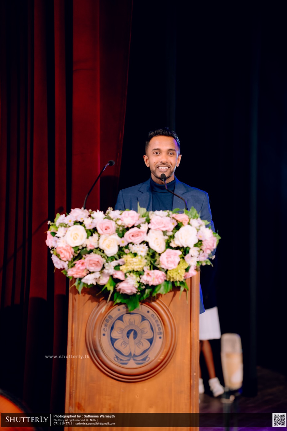 Sampath Soysa speaking at a formal podium with floral arrangement